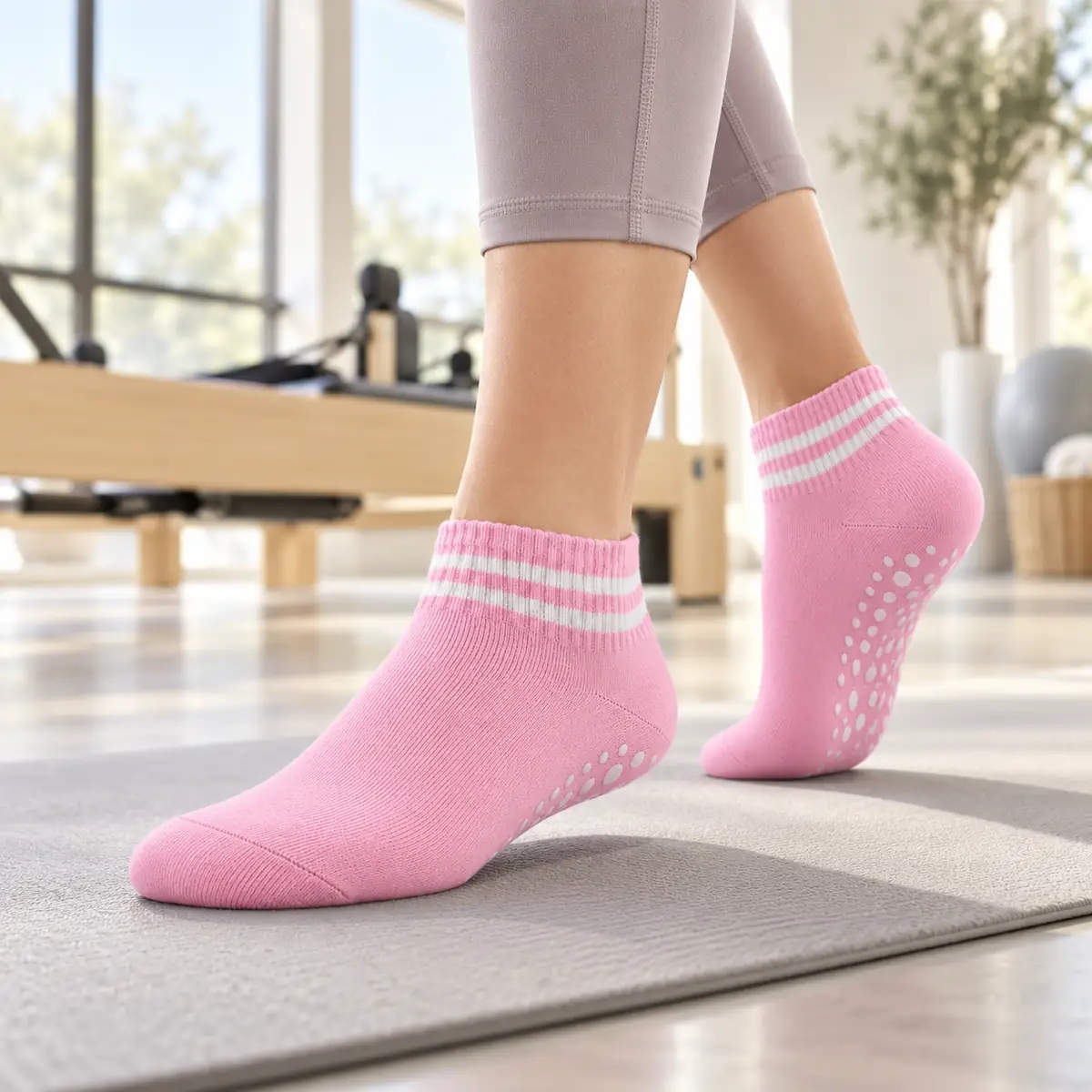 Rosa Knöchel-Socke von Trittfein mit weißen Streifen auf einer grauen Matte, Noppen unter der angehobenen Ferse, helles Studio mit Reformer unscharf im Hintergrund